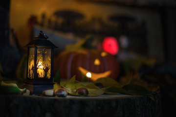 Halloween decorations concept at night. Close up of jack o'lantern, vintage lanterns, pumpkins, skull, autumn leaves. Colorful halloween lights on evening. Happy Halloween scene on wooden background