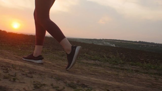 silhouette of young woman legs running on the ground in a field and raising dust, girl in sneakers jogging along a path atsunset, concept of body care and healthy lifestyle