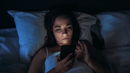 Insomnia and social media addiction concept. Young woman uses smartphone while lying in bed at night