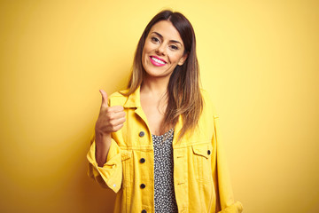 Young beautiful woman wearing denim jacket standing over yellow isolated background doing happy thumbs up gesture with hand. Approving expression looking at the camera with showing success.