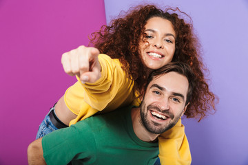 Portrait of excited smiling couple man and woman pointing finger at camera while doing piggyback ride