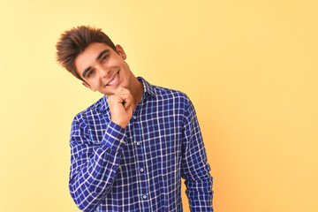 Young handsome man wearing casual shirt standing over isolated yellow background looking confident at the camera smiling with crossed arms and hand raised on chin. Thinking positive.