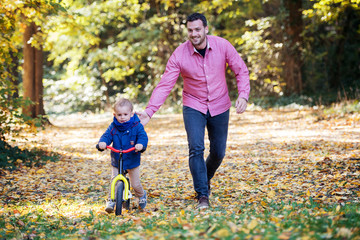 Father and small son with a balance bike on a walk in autumn forest.