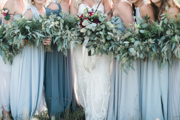 Bride and bridesmaids holding greenery floral arrangements, wearing dresses in shades of blue
