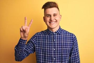 Naklejka premium Young handsome man wearing casual shirt standing over isolated yellow background smiling with happy face winking at the camera doing victory sign. Number two.