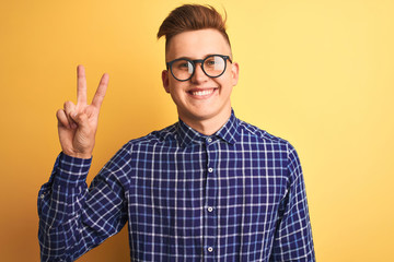 Naklejka premium Young handsome man wearing casual shirt and glasses over isolated yellow background smiling with happy face winking at the camera doing victory sign. Number two.