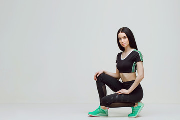 Obraz premium Woman in trendy sportswear relaxing after training. Young beautiful slim brunette girl in fashion black leggings, top and green sneakers posing on gray background.