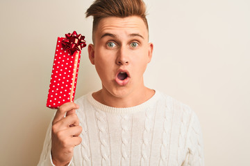 Young handsome man holding valentine gift standing over isolated white background scared in shock...