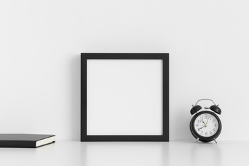 Black square frame mockup with a notebook and a clock on a white table.
