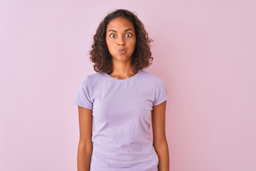 Young brazilian woman wearing t-shirt standing over isolated pink background puffing cheeks with funny face. Mouth inflated with air, crazy expression.