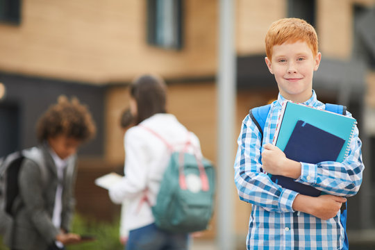 Portrait of red haired schoolboy with books smiling at camera while standing outdoors near the school building