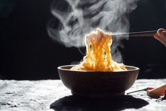 Hand uses chopsticks to pickup tasty noodles with steam and smoke in bowl on wooden background, selective focus. Asian meal on a table, junk food concept