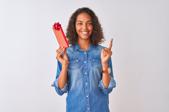 Young brazilian woman holding valentine gift standing over isolated white background surprised with an idea or question pointing finger with happy face, number one