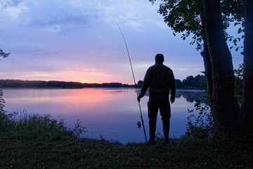 silhouette angler standing on the lake shore during sunrise