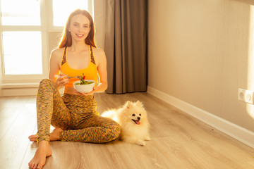 redhaired ginger girl in orange sporty top bra eating salat in evening sun light background and having fun with her white fluffy spitz in big living room