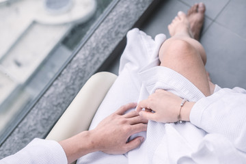 Fototapeta premium Woman sitting on man's laps in white hotel robes touching each other hands. Overhead, man's and woman's legs from above. Intimacy, Valentine's day concept