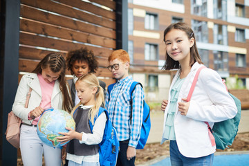 Portrait of Asian schoolgirl with backpack behind her back looking at camera with her classmates looking at globe together in the background outdoors