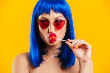 Portrait of glamorous focused young woman wearing blue wig and sunglasses eating lollipop