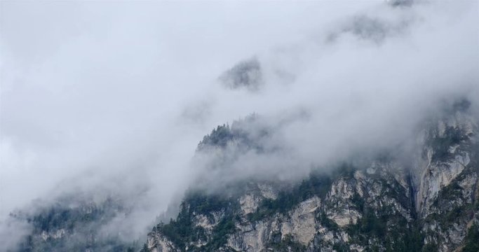 Time lapse of playful white clouds - Stratus fractus silvagenitus (St fra sigen) - on a rock wall with green pine trees in the Austrian Alps after a rainy morning. Hazy and mystical atmosphere.