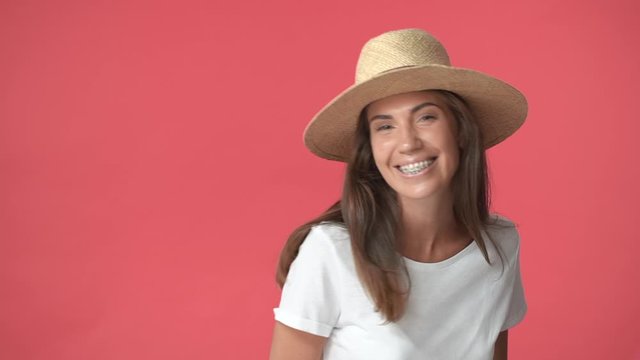 Silly pretty brunette woman in straw hat, white t-shirt standing backwards and turning camera, smiling carefree, enjoying vacation, travelling europe, wearing braces, standing red background