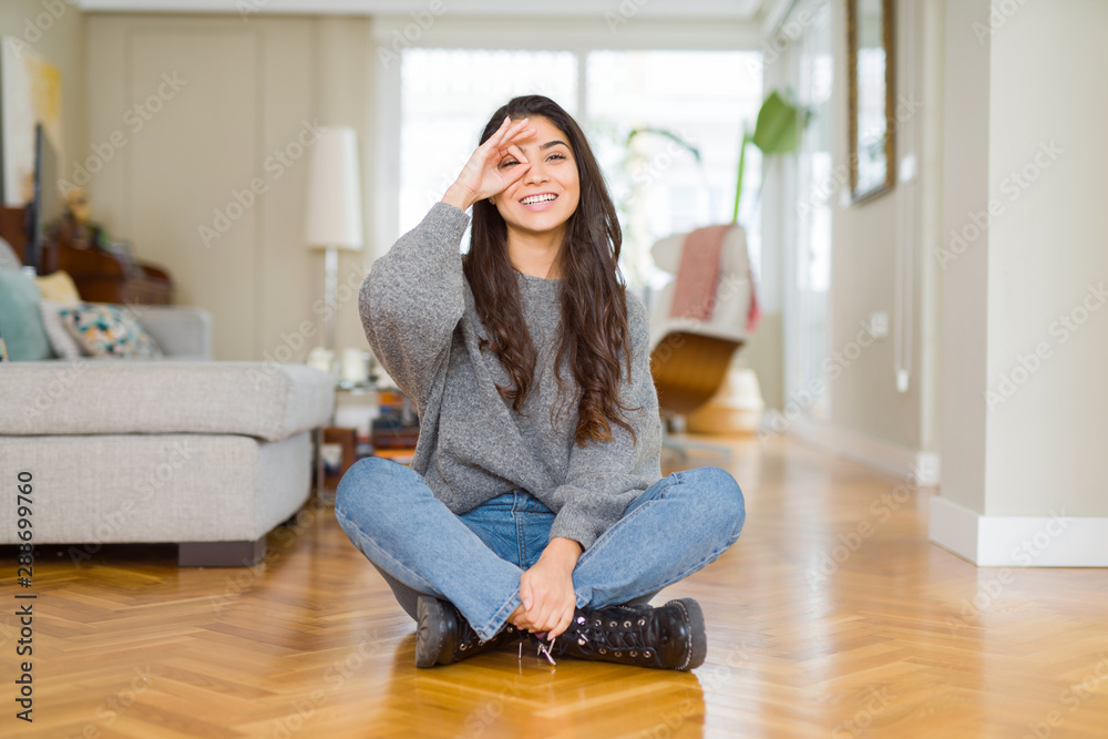 Sticker Young beautiful woman sitting on the floor at home doing ok gesture with hand smiling, eye looking through fingers with happy face.