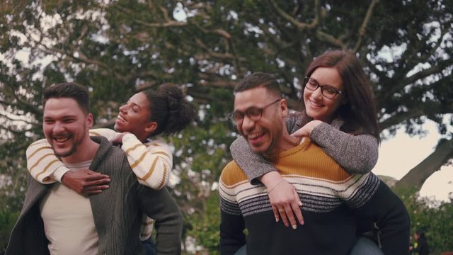 Smiling multi-ethnic couples having fun by carrying their girlfriends on piggyback ride in the park