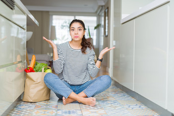 Young woman sitting on the kitchen floor with a paper bag full of fresh groceries clueless and confused expression with arms and hands raised. Doubt concept.