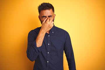 Young handsome indian businessman wearing shirt over isolated yellow background smelling something stinky and disgusting, intolerable smell, holding breath with fingers on nose. Bad smells concept.