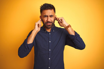 Young handsome indian businessman wearing shirt over isolated yellow background covering ears with fingers with annoyed expression for the noise of loud music. Deaf concept.