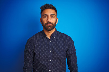 Young indian elegant man wearing shirt standing over isolated blue background with serious expression on face. Simple and natural looking at the camera.