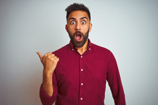 Young indian man wearing red elegant shirt standing over isolated grey background Surprised pointing with hand finger to the side, open mouth amazed expression.
