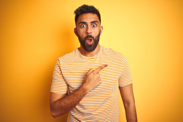 Young indian man wearing t-shirt standing over isolated yellow background Surprised pointing with...