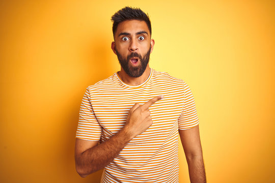 Young indian man wearing t-shirt standing over isolated yellow background Surprised pointing with finger to the side, open mouth amazed expression.