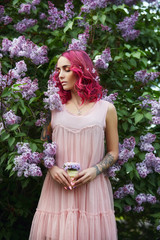 Fashion girl with bright red dyed hair in Apple and lilac flowers. Creative color bright pink, colorist. Woman walks in a Park enjoys the spring. Beautiful tattoos on a woman body
