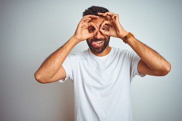 Young indian man wearing t-shirt standing over isolated white background doing ok gesture like binoculars sticking tongue out, eyes looking through fingers. Crazy expression.