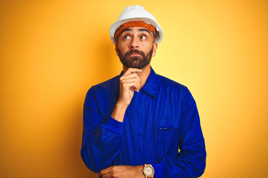 Handsome indian worker man wearing uniform and helmet over isolated yellow background with hand on chin thinking about question, pensive expression. Smiling with thoughtful face. Doubt concept.