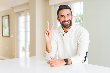 Handsome hispanic man wearing casual white sweater at home showing and pointing up with fingers number two while smiling confident and happy.