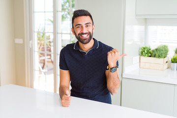 Handsome hispanic man at home smiling with happy face looking and pointing to the side with thumb up.