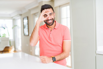 Handsome hispanic man wearing casual t-shirt at home doing ok gesture with hand smiling, eye looking through fingers with happy face.
