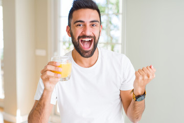 Fototapeta premium Handsome hispanic man drinking healthy orange juice screaming proud and celebrating victory and success very excited, cheering emotion