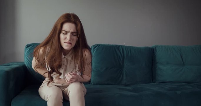 Redhead woman sits on the couch and feels belly pain and anger.