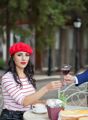 Elegant man in a suit gives a pretty brunette woman in a red cap and striped t-shirt glass of red wine in outdoor cafe