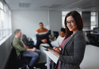 Young smiling businesswoman with tablet computer looking at camera and her creative team working at background