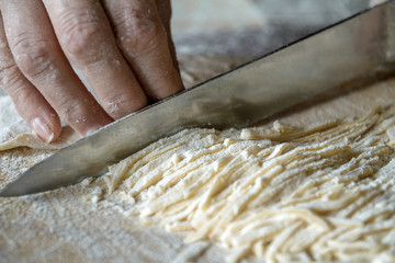Senior woman cuts the dough on wooden board during homemade noodle or pasta production in her home kitchen. Closeup, selective focus