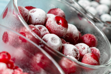 Ripe frozen sweet cherries, red currant and black currant with hoarfrost in the transparent glass cups on blue wooden background. Natural organic healthy food. Closeup, selective focus