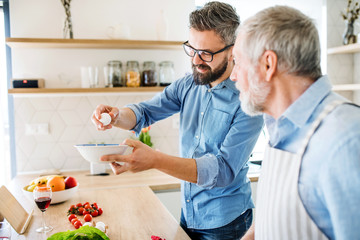 An adult hipster son and senior father indoors at home, cooking.