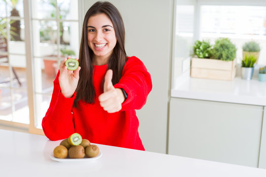 Beautiful young woman eating half fresh green kiwi happy with big smile doing ok sign, thumb up with fingers, excellent sign