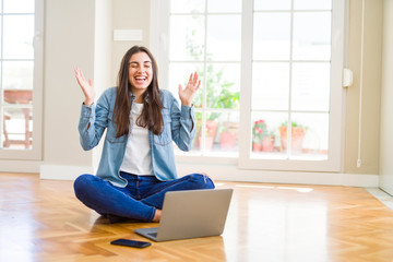 Beautiful young woman sitting on the floor with crossed legs using laptop celebrating mad and crazy for success with arms raised and closed eyes screaming excited. Winner concept