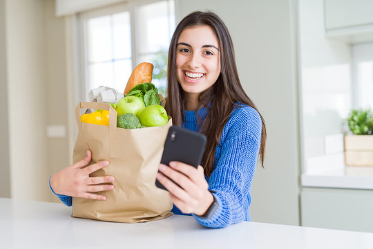 Young woman holding a paper bag full of fresh groceries and using smartphone app for supermarket delivery