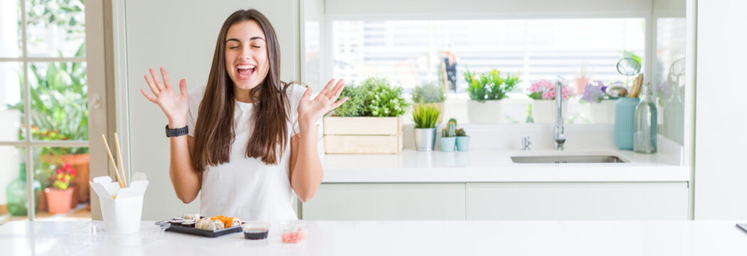 Wide angle picture of beautiful young woman eating asian sushi from delivery celebrating mad and crazy for success with arms raised and closed eyes screaming excited. Winner concept
